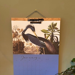 Könnte beinhalten: Wandkalender für Januar 2025 mit einer detaillierten Illustration eines Reiher. Der Kalender wird von einem Holzrahmen und einer schwarzen Klammer gehalten. Der Hintergrund zeigt eine tropische Landschaft.