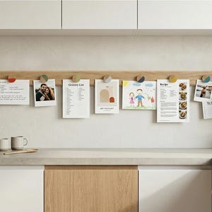 Könnte beinhalten: Eine lange, horizontale Wandleiste aus hellem Holz mit sieben runden Knöpfen mit farbigen Akzenten. Die Leiste hält verschiedene Papiere, darunter eine Einkaufsliste, ein Rezept, eine Kinderzeichnung und mehrere Fotografien.