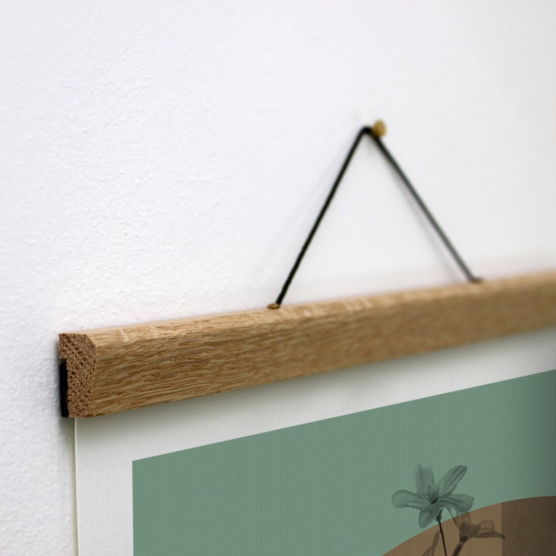 May include: A wooden poster hanger with a black string and brass hardware. The hanger is attached to a white wall and is holding a poster with a green background and a flower.