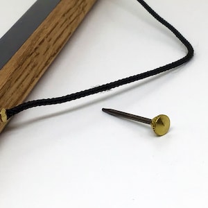May include: A close-up of a wooden frame with a black cord attached to a brass nail. The nail is on a white surface.