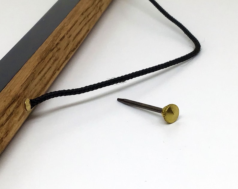 May include: A close-up of a wooden frame with a black cord attached to a brass nail. The nail is on a white surface.