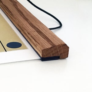 May include: Close-up of a wooden poster hanger with a black hanging cord and a black magnetic strip. The poster hanger is made of natural wood with a light brown finish.