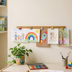 May include: A horizontal wooden wall-mounted display rail featuring five circular pegs with colourful painted accents. The rail holds several children's drawings, including a family portrait, a rainbow, a rocket ship, a cat, and a tree.