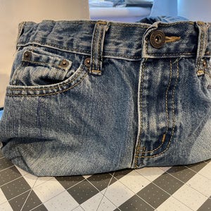 Może przedstawiać: Jeansowa kopert&oacute;wka wykonana z przetworzonego niebieskiego denimu. Torba posiada oryginalne kieszenie, szlufki i zapięcie na guzik. Unikalny, ekologiczny dodatek.