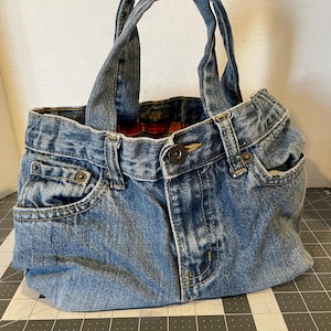 Może przedstawiać: Ręcznie wykonana torba typu tote z denimu wykonana z przetworzonych niebieskich dżins&oacute;w. Torba posiada oryginalne kieszenie dżinsowe, zapięcie na guziki i szlufki na pasek, ze stabilnymi uchwytami z denimu. Wnętrze wyłożone jest czerwoną i czarną kratą.
