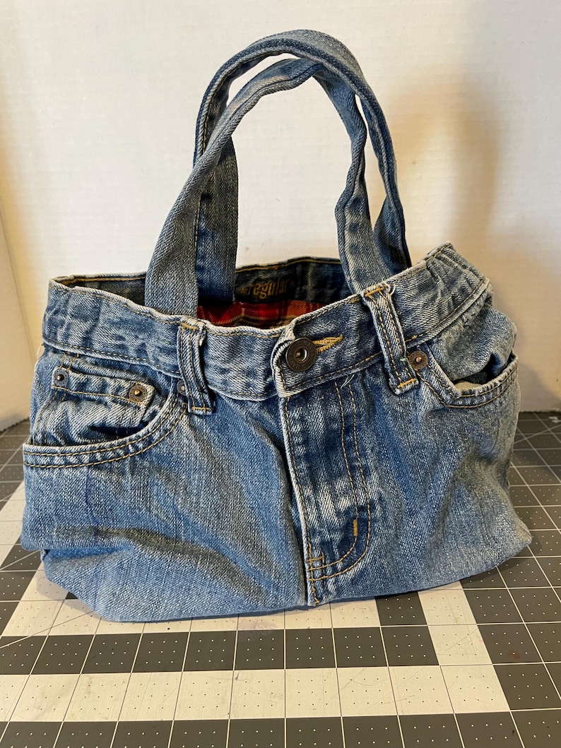 Może przedstawiać: Ręcznie wykonana torba typu tote z denimu wykonana z przetworzonych niebieskich dżins&oacute;w. Torba posiada oryginalne kieszenie dżinsowe, zapięcie na guziki i szlufki na pasek, ze stabilnymi uchwytami z denimu. Wnętrze wyłożone jest czerwoną i czarną kratą.