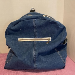 May include: A denim backpack with a curved top and a white zipper. The backpack has a black metal ring at the top and a white stripe along the top seam. The bag is made of blue denim.