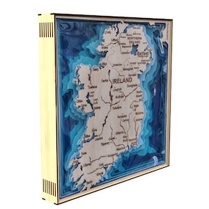 Może przedstawiać: Trójwymiarowa drewniana mapa Irlandii z niebieskimi, warstwowymi szczegółami oceanu. Mapa jest oprawiona w jasną drewnianą ramę i zawiera tekst "IRELAND" oraz główne miasta.