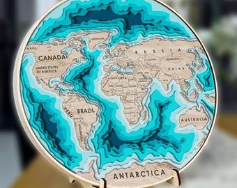 May include: A circular wooden world map wall art piece featuring layered, laser-cut continents in a light wood finish set against deep, multi-toned blue ocean depths. The map includes clearly engraved country names and rests on a small stand.