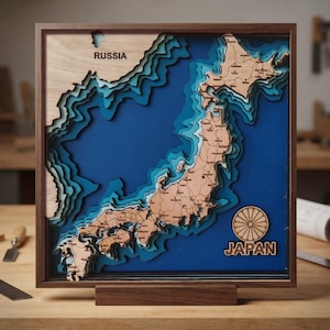 May include: A square wooden wall art piece featuring a layered 3D bathymetric map of Japan and parts of Russia. The map uses light wood for landmasses and various shades of blue for ocean depths, encased in a dark wood frame with the text JAPAN.