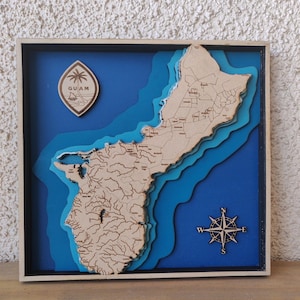 May include: A wooden layered map of Guam, featuring detailed coastlines and topography. The map is set against a blue background, with a compass rose and a Guam emblem. The frame is black and natural wood.