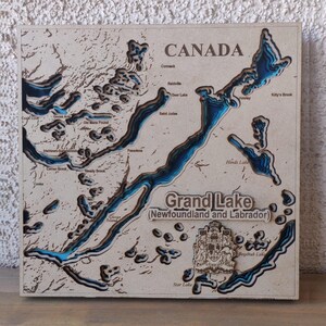 May include: A wooden map of Grand Lake in Newfoundland and Labrador, Canada. The map features a detailed, layered design with blue water and engraved place names. The word "CANADA" is at the top.