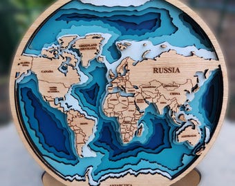May include: A circular wooden world map wall art piece featuring layered, laser-cut continents in natural wood tones set against a multi-tonal blue ocean background. The map includes engraved country names and labels like Russia, Canada, and Antarctica.