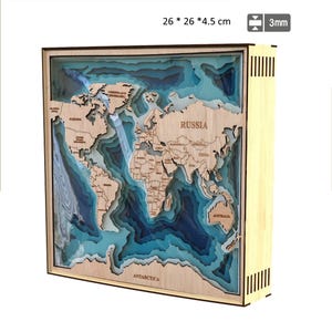 Może przedstawiać: Drewniana mapa świata, 26 x 26 x 4,5 cm, z warstwami niebieskiego i przezroczystego akrylu reprezentującymi oceany. Kontynenty wycięte są z jasnego drewna i opisane. Rama jest z jasnego drewna z ciemną drewnianą obwódką.