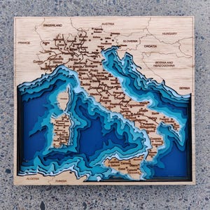 Puede incluir: Un mapa de madera multicapa de Italia, cortado con láser para mostrar detalles topográficos. El mapa muestra la forma de Italia con tonos azules para el agua y la tierra. Los nombres de las ciudades y las fronteras están etiquetados, en un marco de madera natural.