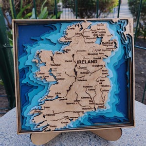Puede incluir: Un mapa de madera en capas de Irlanda, con nombres de ciudades y contornos geográficos. El mapa está enmarcado en madera oscura y sobre un fondo azul, creando un efecto 3D. El mapa mide aproximadamente 25 cm cuadrados.