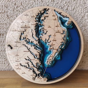 May include: A round, wooden topographic map of the Chesapeake Bay area, including Washington D.C. and surrounding cities. The map features layered, laser-cut wood with blue resin representing water, and city names etched into the wood.