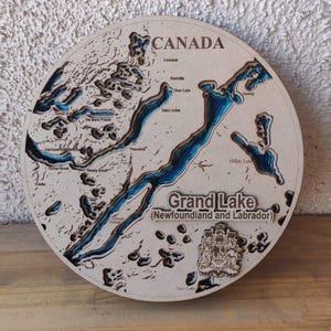 May include: A round, wooden map of Grand Lake in Newfoundland and Labrador, Canada. The map features laser-cut layers showing the lake's depth in blue, with the names of surrounding areas and the word "CANADA" at the top.