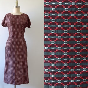 May include: A vintage, knee-length dress with short sleeves, displayed on a mannequin. The dress features a repeating pattern of red, white, and black squares. The fabric's texture is visible. A close-up of the fabric shows the pattern in detail.