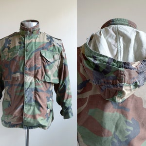 May include: A green, brown, and black camouflage jacket with a hood. The jacket has a button-up front and two large pockets on the front.