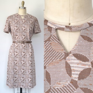 May include: A vintage knee-length dress in shades of brown and beige, featuring a geometric pattern. The dress has short sleeves, a keyhole neckline, and a brown belt cinching the waist. The dress is displayed on a mannequin.