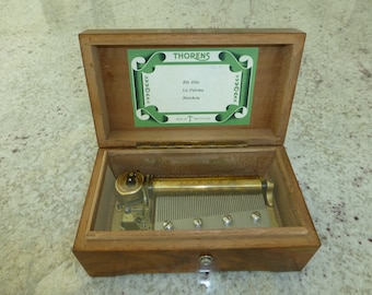May include: A wooden music box with a glass lid. The music box is open and the mechanism is visible. The music box plays the songs "Rio Rita", "La Paloma", and "Marcheta". The music box is made in Switzerland.