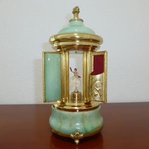 May include: A decorative music box featuring a ballerina figurine. The box is light green and gold, with a domed top and intricate detailing. The ballerina wears a white tutu and has a red arm raised.