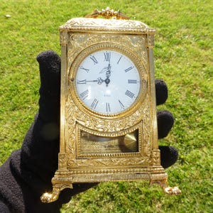 May include: A gold-toned mantel clock with ornate details and a white face with black Roman numeral hour markers. The clock has a pendulum and a glass front.