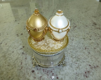 May include: Two decorative eggs, one gold and one white, with pearl and gold embellishments, sit atop a silver music box with a gold trim.