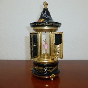 May include: A decorative music box with a black and gold finish. The music box features a ballerina figurine in a pink dress. The box has ornate detailing and a decorative top with a painted design. The doors are open, revealing the ballerina.