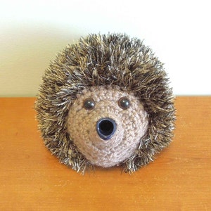 May include: A crocheted hedgehog ornament with a surprised expression. The hedgehog has a brown body, large black eyes, and a dark blue mouth. Its spiky fur is a mix of brown and silver yarn. It rests on a wooden surface.