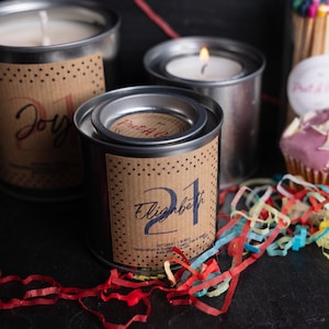 Puede incluir: Tres velas en latas de metal con etiquetas de papel marrón. Las velas están etiquetadas como "Joy", "Pret-a-Go" y "Elizabeth 21". En el fondo, hay un cupcake rosa con virutas de chocolate blanco y confeti de colores.