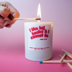 May include: A white candle with the text "I like big books & I cannot lie BOOK CLUB" in pink. A match is lit, igniting the candle's wick. The candle is labeled "Lavender & Vanilla Soy Candle 35 ml 200g".