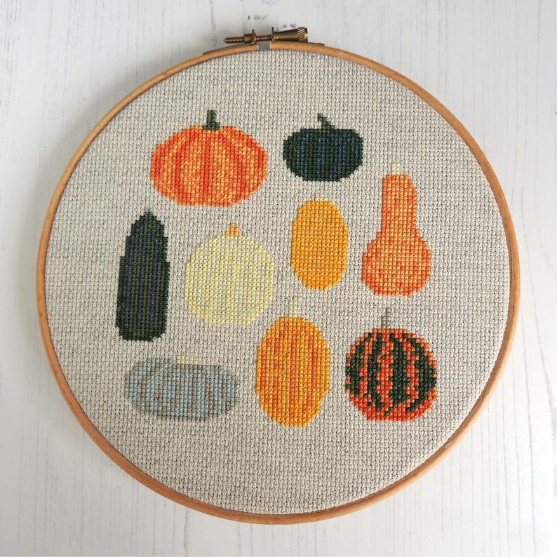 K&ouml;nnte beinhalten: Ein Kreuzstich-Stickrahmen mit verschiedenen bunten K&uuml;rbissen, Flaschenk&uuml;rbissen und K&uuml;rbissen. Die Stickerei ist auf einem beigefarbenen Stoff.