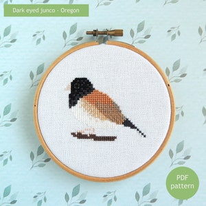 May include: Cross stitch pattern for a dark-eyed junco, a small brown and white bird with a black head, in a wooden embroidery hoop. The pattern is titled "Dark eyed junco - Oregon".