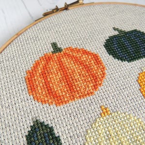 K&ouml;nnte beinhalten: Nahaufnahme eines Kreuzstich-Stickrahmens mit einem Design aus drei K&uuml;rbissen in verschiedenen Farben: orange, gr&uuml;n und gelb. Die K&uuml;rbisse sind mit verschiedenen Farbt&ouml;nen von orangem, gr&uuml;nem und gelbem Garn gestickt.