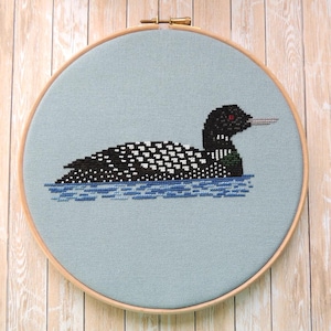 Puede incluir: Una bordado de punto de cruz de un colimbo nadando en agua azul, enmarcado en un aro de madera claro.