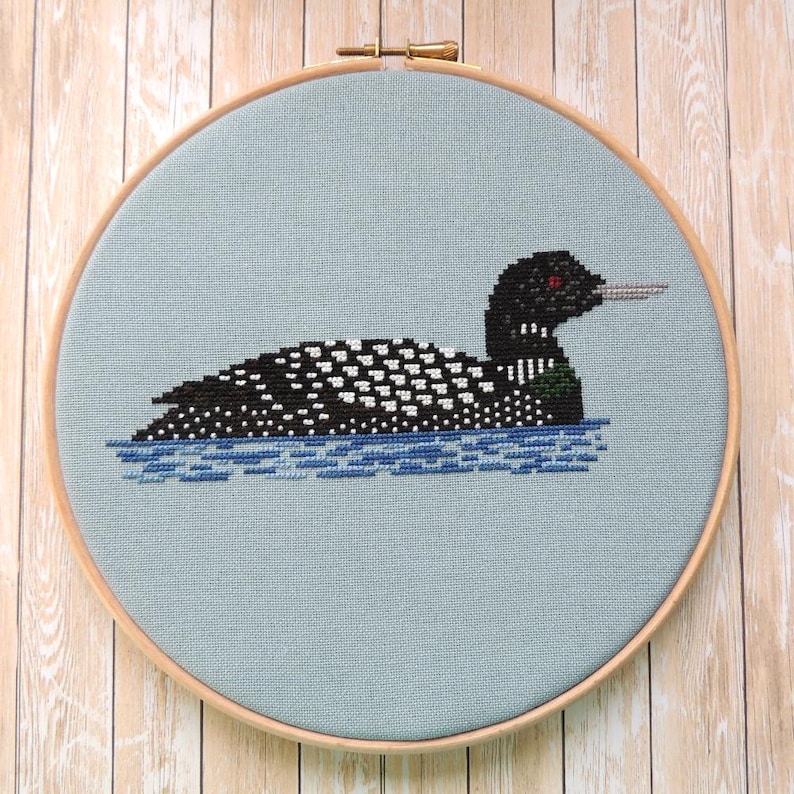 May include: A cross-stitch embroidery of a loon swimming in blue water, framed in a light wood hoop.
