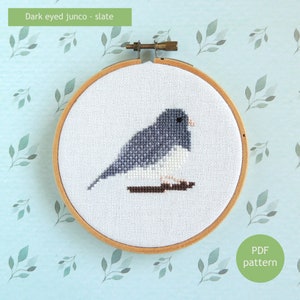 Può includere: Uno schema di ricamo a punto croce di un junco dagli occhi scuri color ardesia su uno sfondo bianco. L'uccello è posato su un ramo. Lo schema è in un telaio da ricamo in legno. Il testo "PDF pattern" è su un cerchio verde nell'angolo in basso a destra.