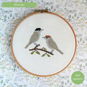 May include: Cross stitch pattern of two blackcap birds perched on a branch. The pattern is in a wooden embroidery hoop. The text "Blackcaps" is at the top of the image and "PDF pattern" is at the bottom.