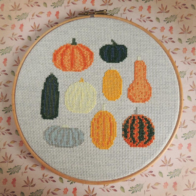 K&ouml;nnte beinhalten: Ein Kreuzstich-Stickrahmen mit einer Auswahl an Herbstk&uuml;rbissen und -k&uuml;rbissen in verschiedenen Farben und Formen. Die K&uuml;rbisse und K&uuml;rbisse sind in einem einfachen, modernen Stil gestickt.