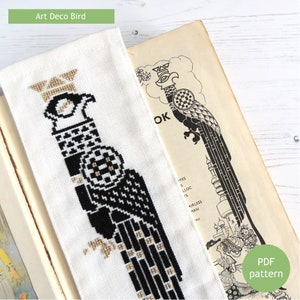 May include: A black and gold cross-stitch pattern of an Art Deco bird on white Aida cloth. The pattern is displayed on a book with a similar design.