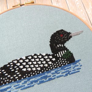 May include: A black and white loon cross-stitch pattern on a light blue fabric. The loon is swimming in blue water.