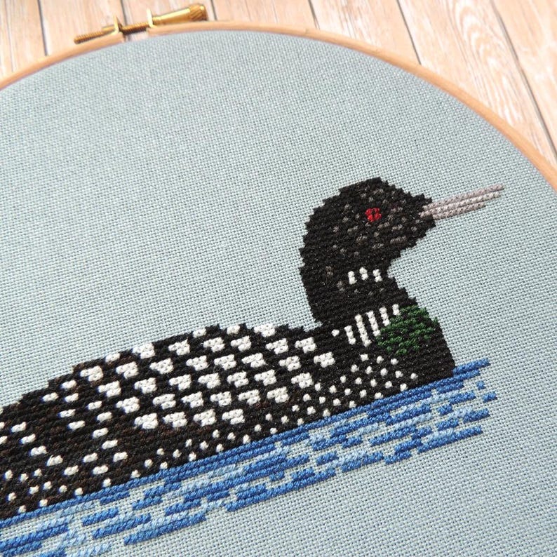 May include: A black and white loon cross-stitch pattern on a light blue fabric. The loon is swimming in blue water.