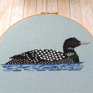 May include: Cross-stitch embroidery of a loon swimming in blue water on a light blue background. The loon is black with white spots and has a red eye.