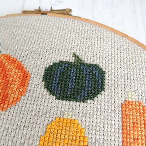 K&ouml;nnte beinhalten: Nahaufnahme einer Kreuzstich-Stickerei mit verschiedenen K&uuml;rbissen in Orange-, Gelb- und Gr&uuml;nt&ouml;nen. Die K&uuml;rbisse sind auf einem wei&szlig;en Stoff mit subtiler Textur gestickt.