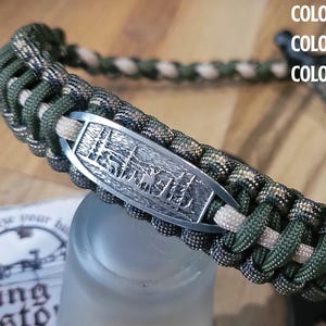May include: A forest camo, olive green, and tan paracord lanyard with a metal emblem featuring a forest scene. The lanyard is braided and has a black clasp. The text on the image reads "COLOR #1: FOREST CAMO, COLOR #2: OLIVE GREEN, COLOR #3: TAN."