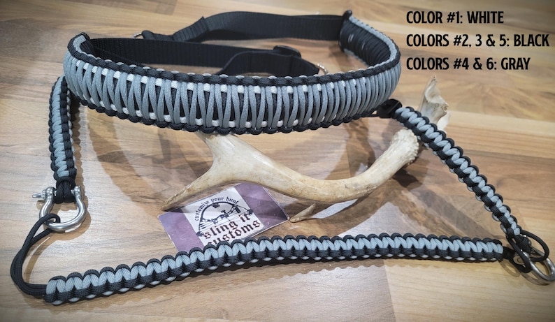 May include: A black, white, and grey paracord sling with metal fixings. The sling is shown with a deer antler and a tag that reads "Bling it Customs". The colour scheme includes white, black, and grey.
