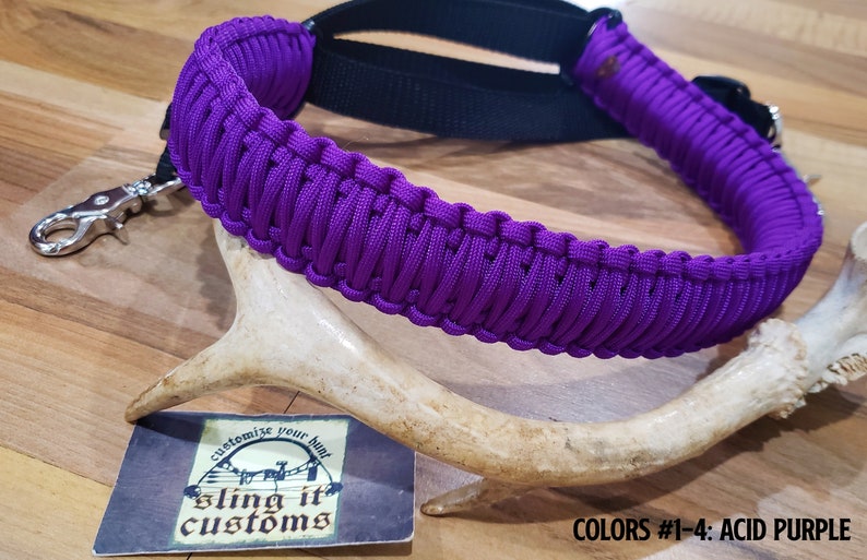 May include: Purple paracord sling with a black strap and a silver hook. The sling is made with a braided pattern and is labeled "sling it customs". The text "COLORS #1-4: ACID PURPLE" is visible at the bottom of the image.