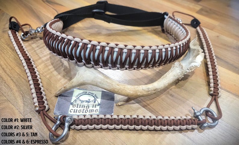 May include: A brown and beige paracord rifle sling with silver-coloured metal fixings. The sling is made of woven cord and has a padded neck strap. The image also shows a deer antler and a business card with the text "Bling it Customs".